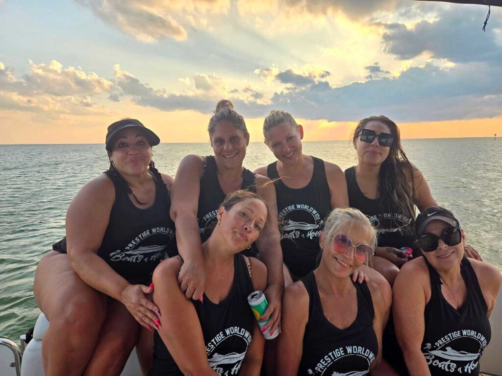 Group of women in black tank tops with "Prestige Worldwide Boats & Hoes" logo, enjoying a sunset on a boat.