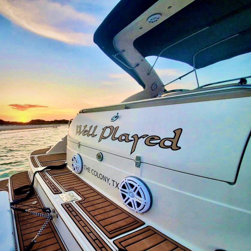 Boat stern with name "Well Played" and "The Colony, TX," shown at sunset near the shore.