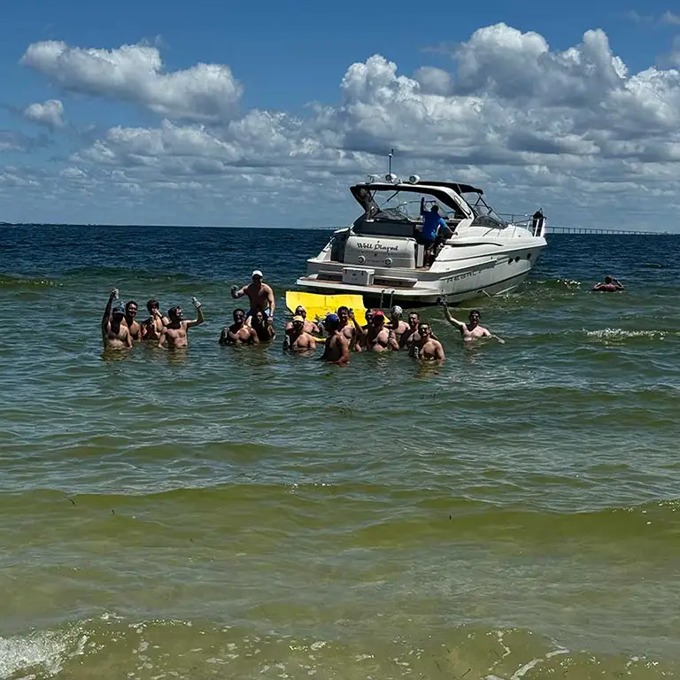 A group of people standing in shallow water near a boat, enjoying a sunny day with clear skies.