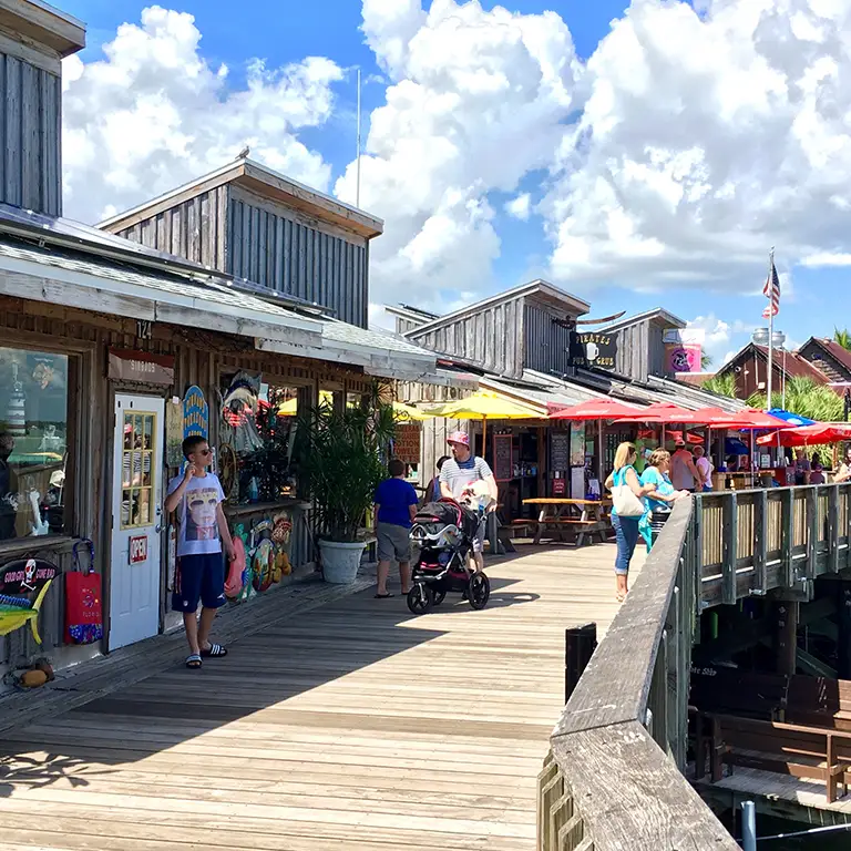 People strolling along a wooden boardwalk lined with shops and colorful umbrellas on a sunny day.