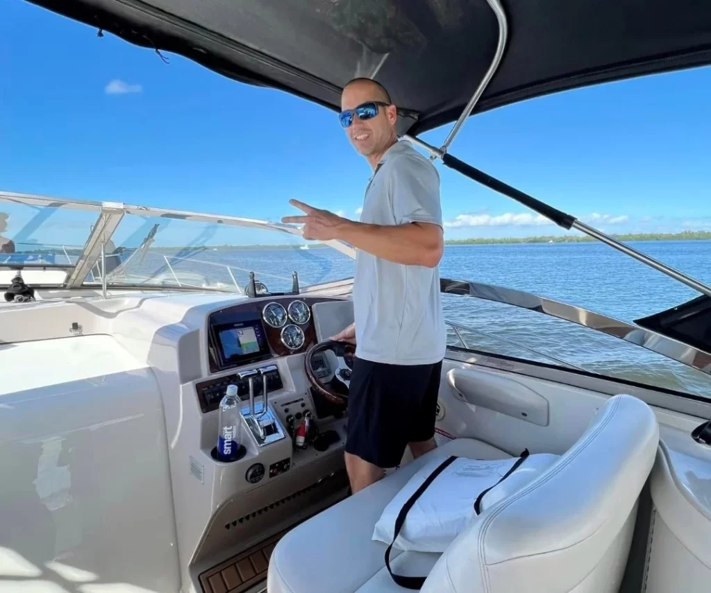 Smiling man steering a yacht on open water, giving a peace sign under a clear blue sky.