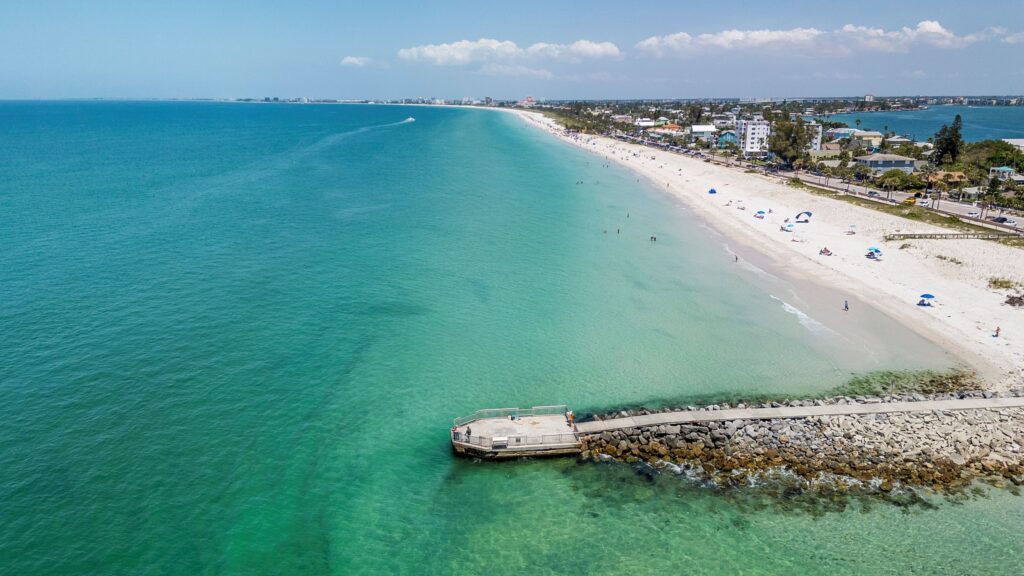 Aerial view of a long sandy beach with turquoise water, small pier, and scattered beachgoers.