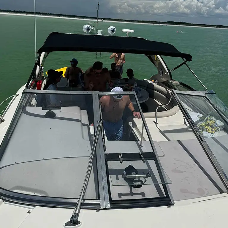 A group of people relaxing on a boat anchored in calm, greenish water near a sandy shoreline.