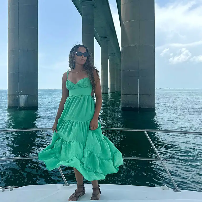 Woman in a flowing green dress stands on a boat beneath a bridge, looking away from the camera.