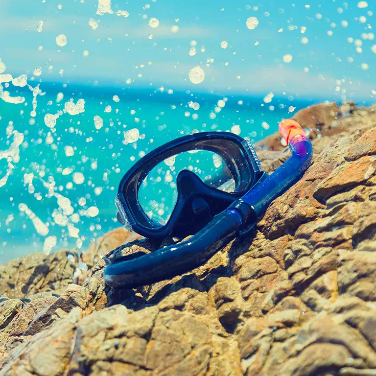 Snorkel mask and tube resting on rocks by the ocean, with splashing water and turquoise sea in the background.