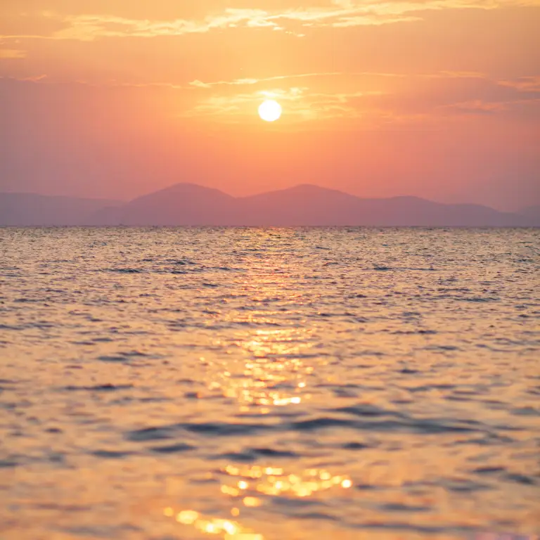 Sunset over calm ocean waters with a warm glow reflecting, and mountains in the distant horizon.