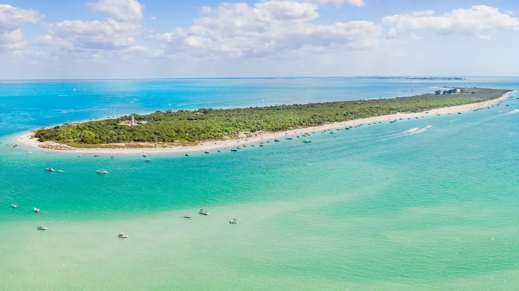 Aerial view of a long, green island with boats anchored around in turquoise waters.