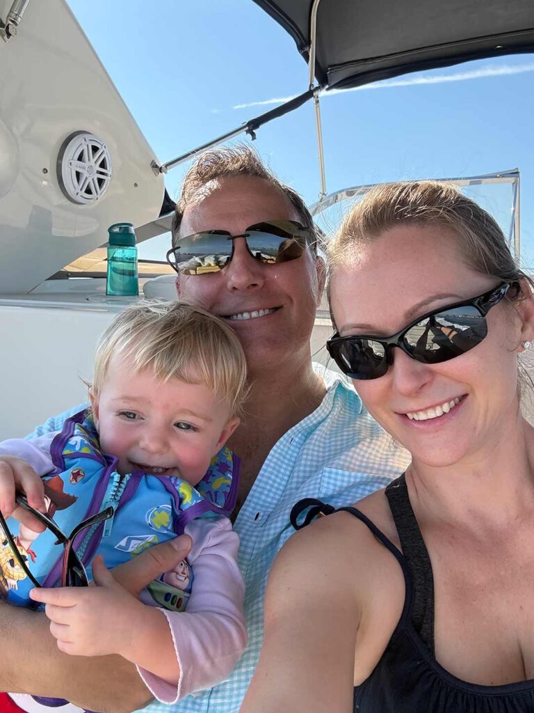 Family taking a selfie on a boat; toddler in a life vest smiles while held by an adult.