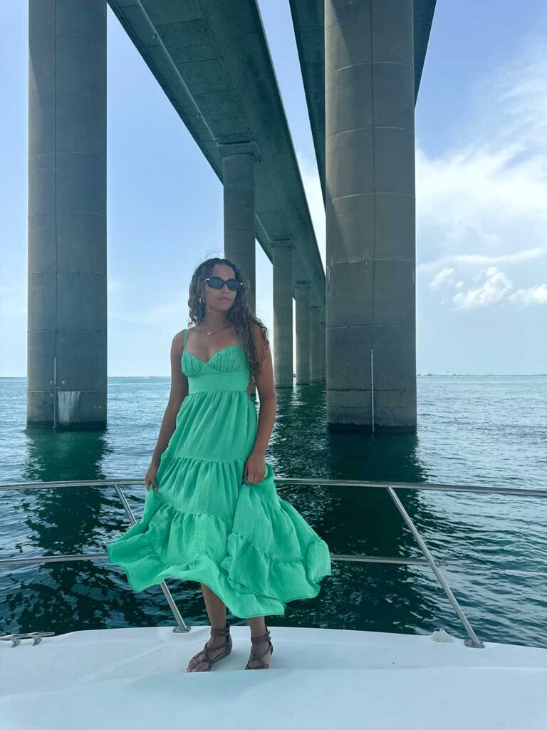 Woman in a flowing green dress stands on a boat beneath a bridge, looking away from the camera.