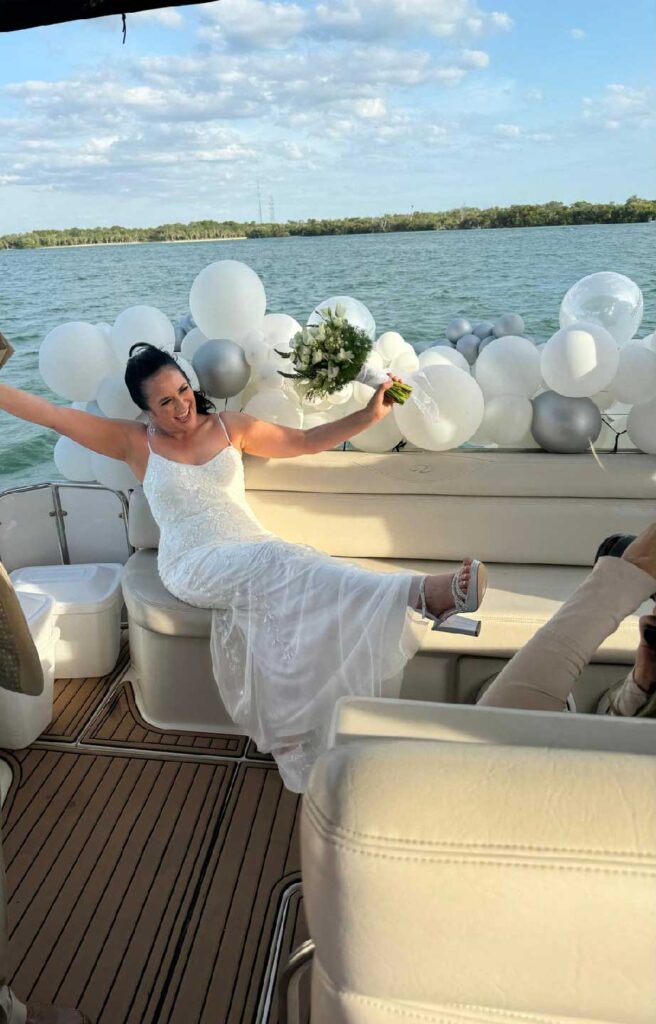 Bride in white dress posing joyfully on a boat decorated with white and silver balloons.