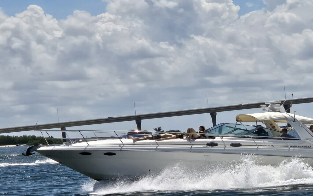 People relaxing on a yacht cruising through open water on a partly cloudy day near a bridge.