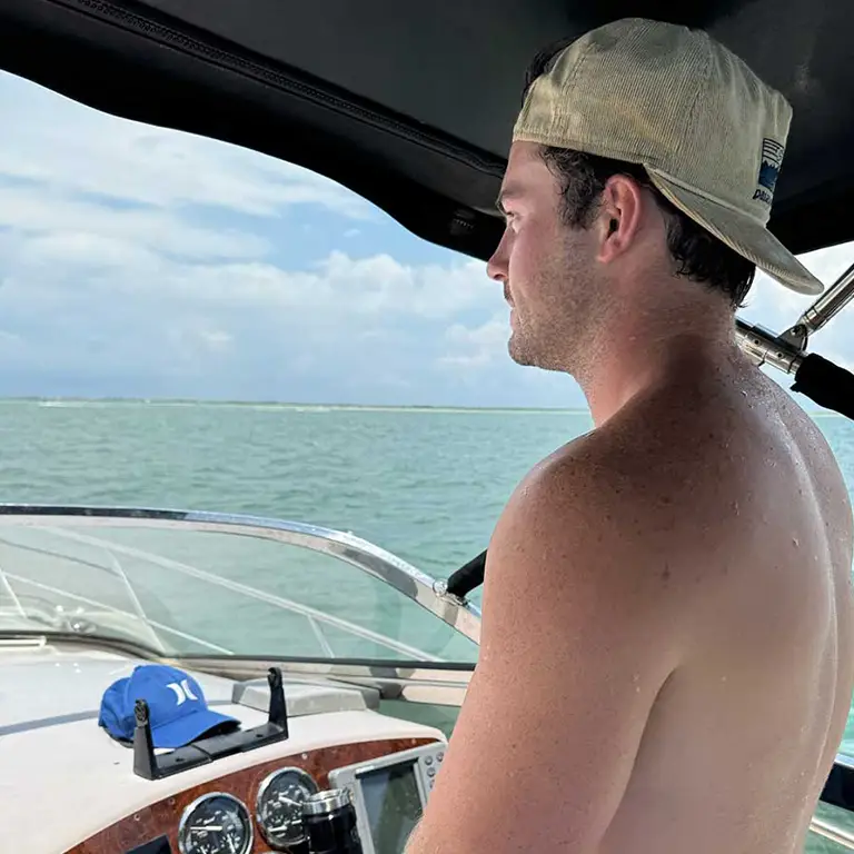 Man in a backward cap steering a boat, gazing at the water on a sunny day.